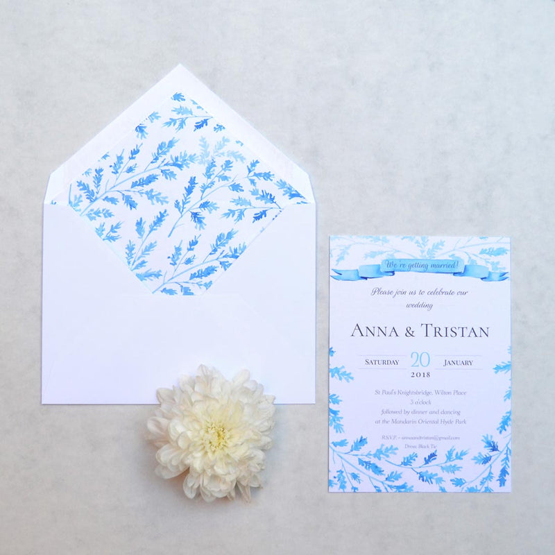 A blue ribbon banner and a frame of winter foliage sprigs adorn the Lillehammer Wedding Invitation and paper lining of the envelope
