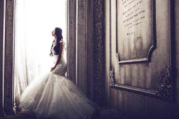 A bride in a flowing wedding dress stands beside a veiled window