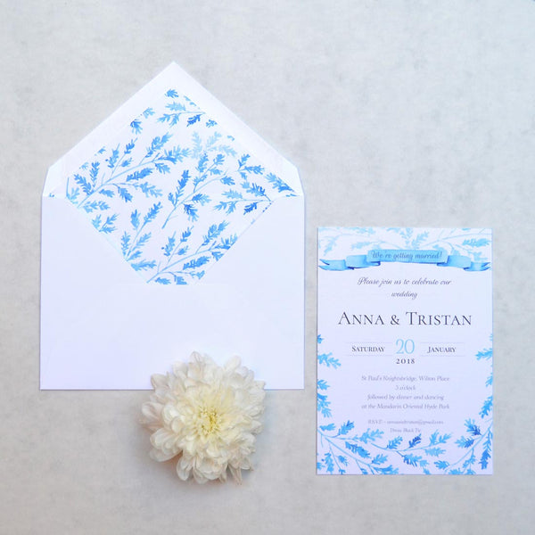 A blue ribbon banner and a frame of winter foliage sprigs adorn the Lillehammer Wedding Invitation and paper lining of the envelope